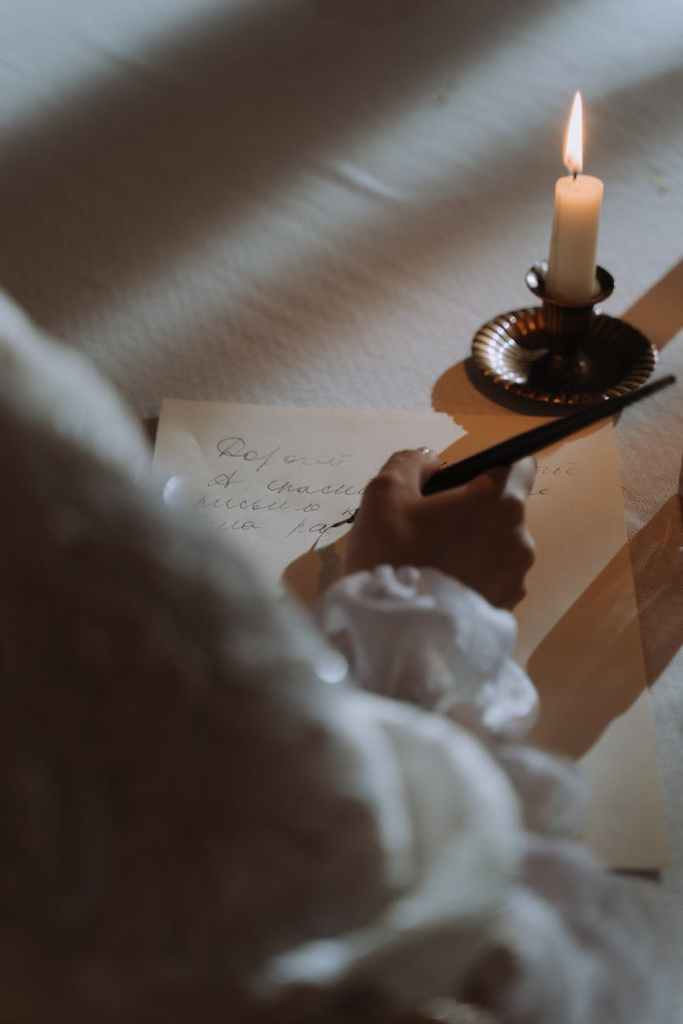 A hand in a frilly shirt writing a letter by candle light using a fountain pen.